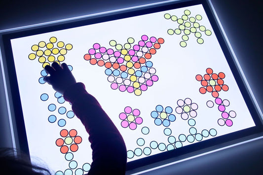 Uso de formas coloridas e translúcidas sobre a prancheta iluminada, proporcionando um ambiente visualmente estimulante e adequado para aprendizado sensorial.